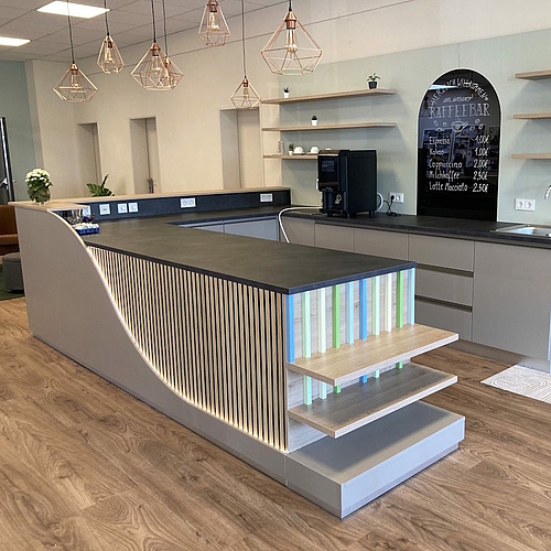 Moderner Thekenbereich mit geschwungener Front, vertikalen Holzleisten, integrierter Beleuchtung und offenen Regalböden im Hintergrund.